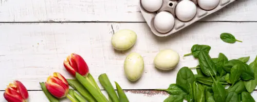 Eierkarton mit weißen Eiern neben frischem Spinat, Tulpen und grünen Eiern auf hellem Holztisch.