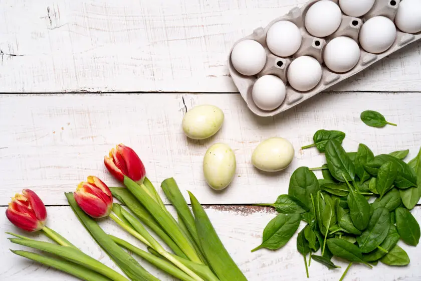 Eierkarton mit weißen Eiern neben frischem Spinat, Tulpen und grünen Eiern auf hellem Holztisch.