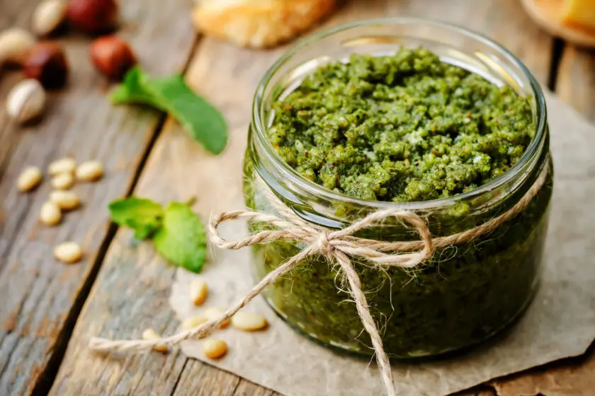 Glas mit grünem Basilikum Pesto, garniert mit Kordel, daneben Pinienkerne auf Holztisch.