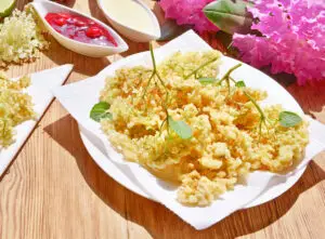 Ausgebackene Holunderblüten auf Teller mit fruchtigen Beilagen serviert.